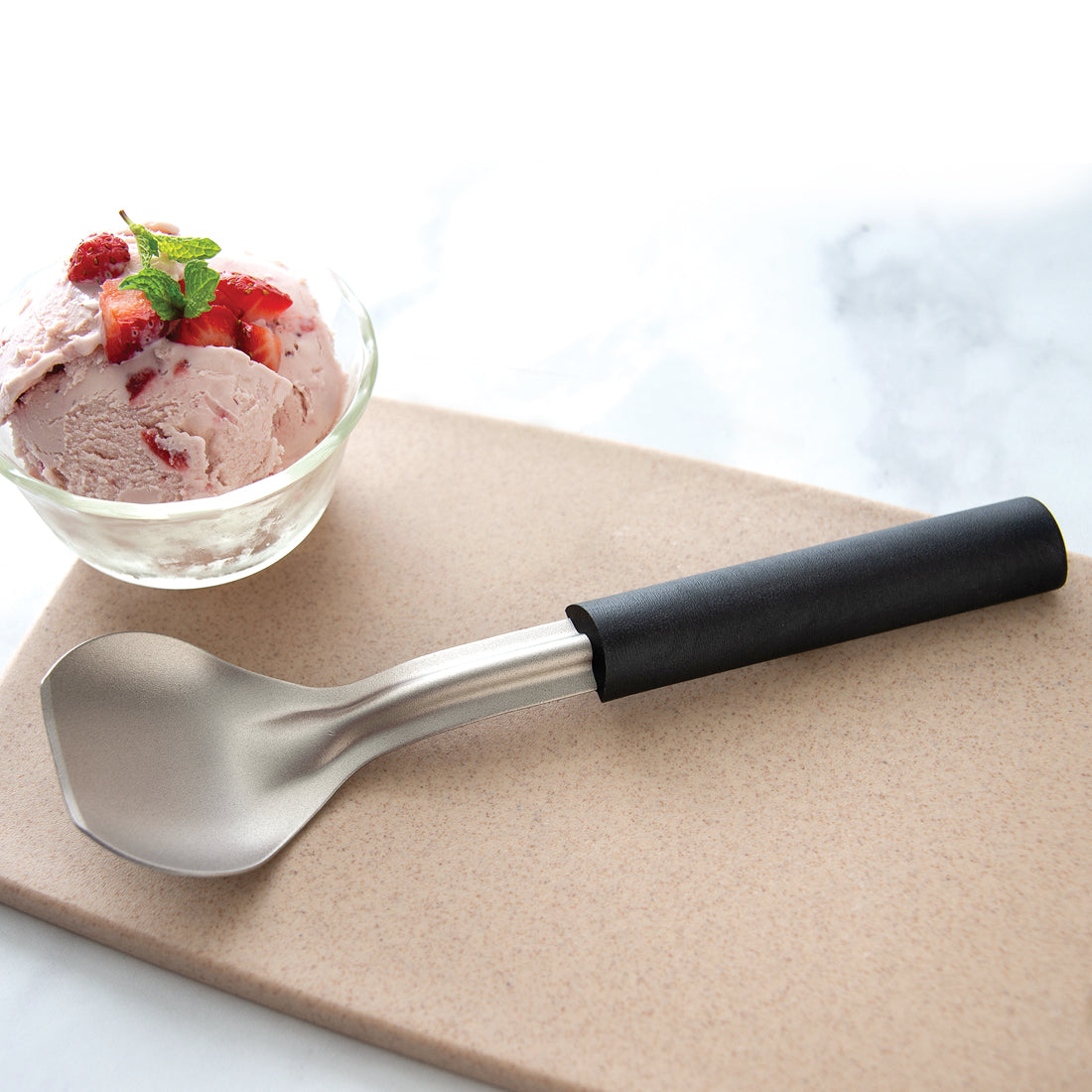 A fresh dipped serving of strawberry ice cream in a glass bowl garnished with fresh mint alongside the Rada Ice Cream Scoop.