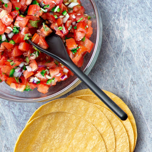 A Rada Mixing Spoon in a bowl of tomatoes, greens, and onions next to taco shells on a light colored background.