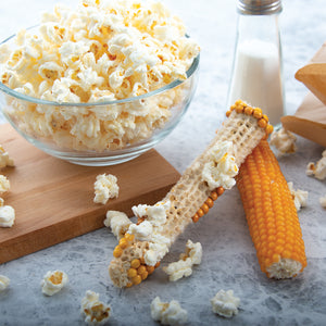 An ear of just popped popcorn next to an ear of unpopped popcorn. A bowl of popcorn sitting on a cutting  board. 