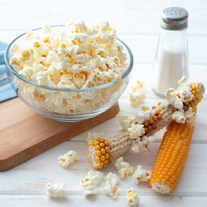 An ear of just popped popcorn next to an ear of unpopped popcorn. A bowl of popcorn sitting on a cutting board.