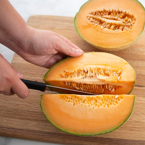 Black handled slicer cutting into a cantaloupe on a wooden cutting board.