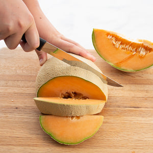 Black handled slicer cutting into a cantaloupe on a wooden surface.