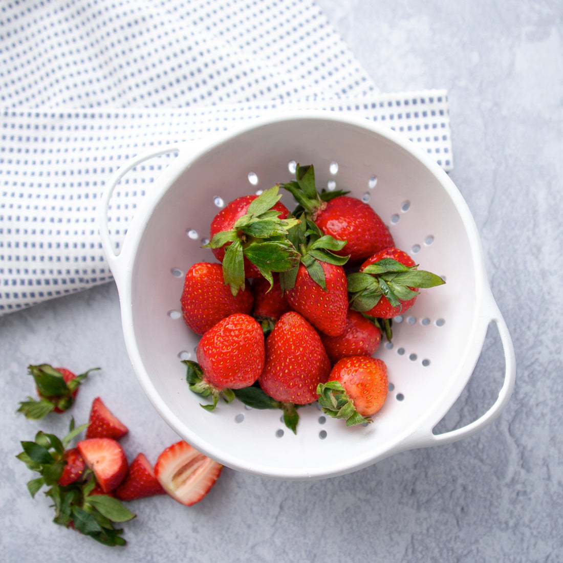 White colander full of strawberries next to a blue and white dishcloth.