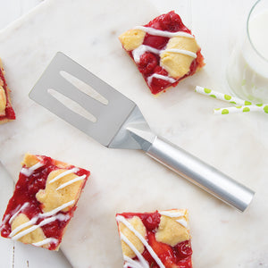 A Rada Cutlery spatula on a marbled surface with squares of cherry bars and a glass of milk with green polka-dot straws.