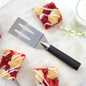 A black resin spatula on a marbled surface with squares of cherry bars and a glass of milk with green polka-dot straws. 