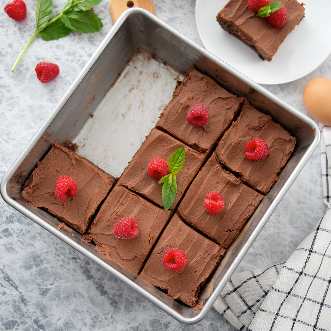 A square baking pan with frosted chocolate brownies topped with raspberries. White serving plate.
