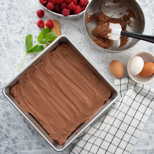 A pan of chocolate frosted brownies next to a bowl of frosting, a spatula, and a bowl of berries. 
