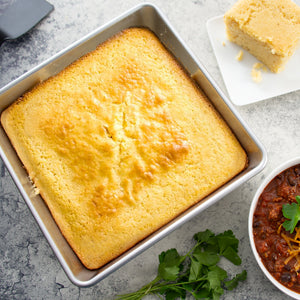 A Rada 9-inch baking pan with unsliced cornbread beside cilantro and a bowl of black bean chili. 