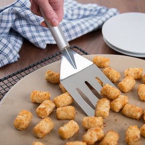 Turnover spatula with silver handle scooping up tater tots from a baking stone. 