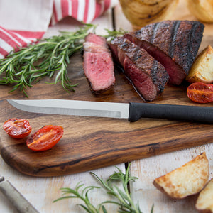 A sliced steak on a cutting board with rosemary, grilled potatoes and tomatoes and a Rada Cutlery utility/steak knife. 