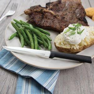 The utility/steak knife on a dinner plate with green beans, a baked potato with sour cream and chives and a T-bone steak.